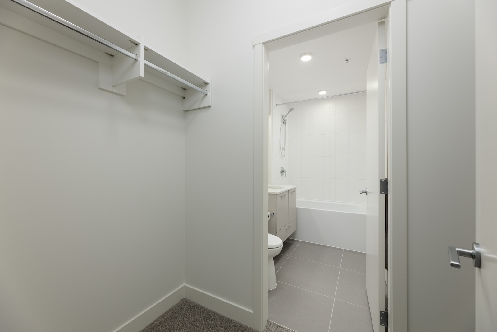 Empty closet space with an open door leading to a bathroom featuring a bathtub, toilet, and vanity, under ceiling lights.