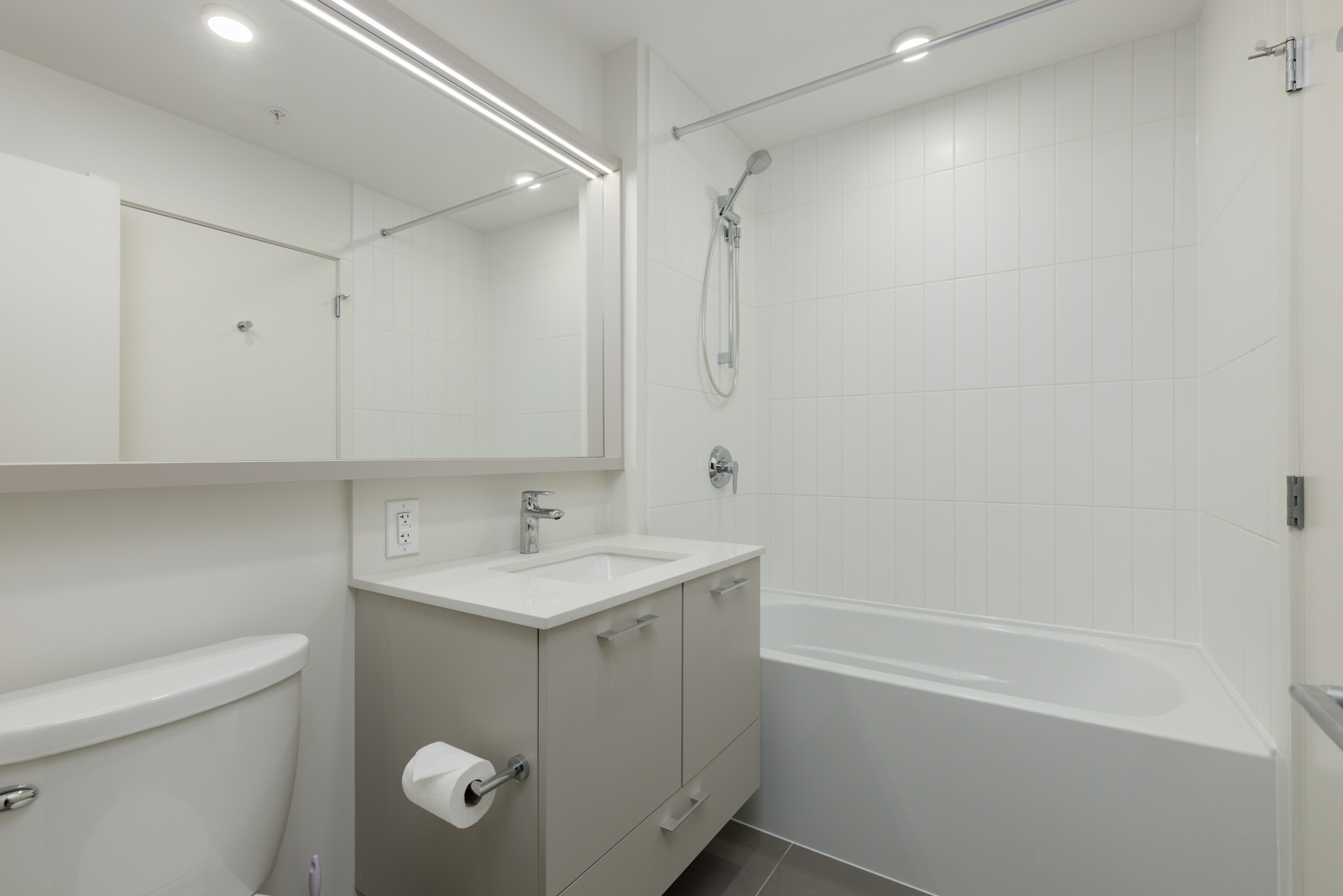 Modern bathroom with light gray vanity, rectangular mirror, toilet, and white bathtub with overhead shower. Walls and backsplash are tiled in white.
