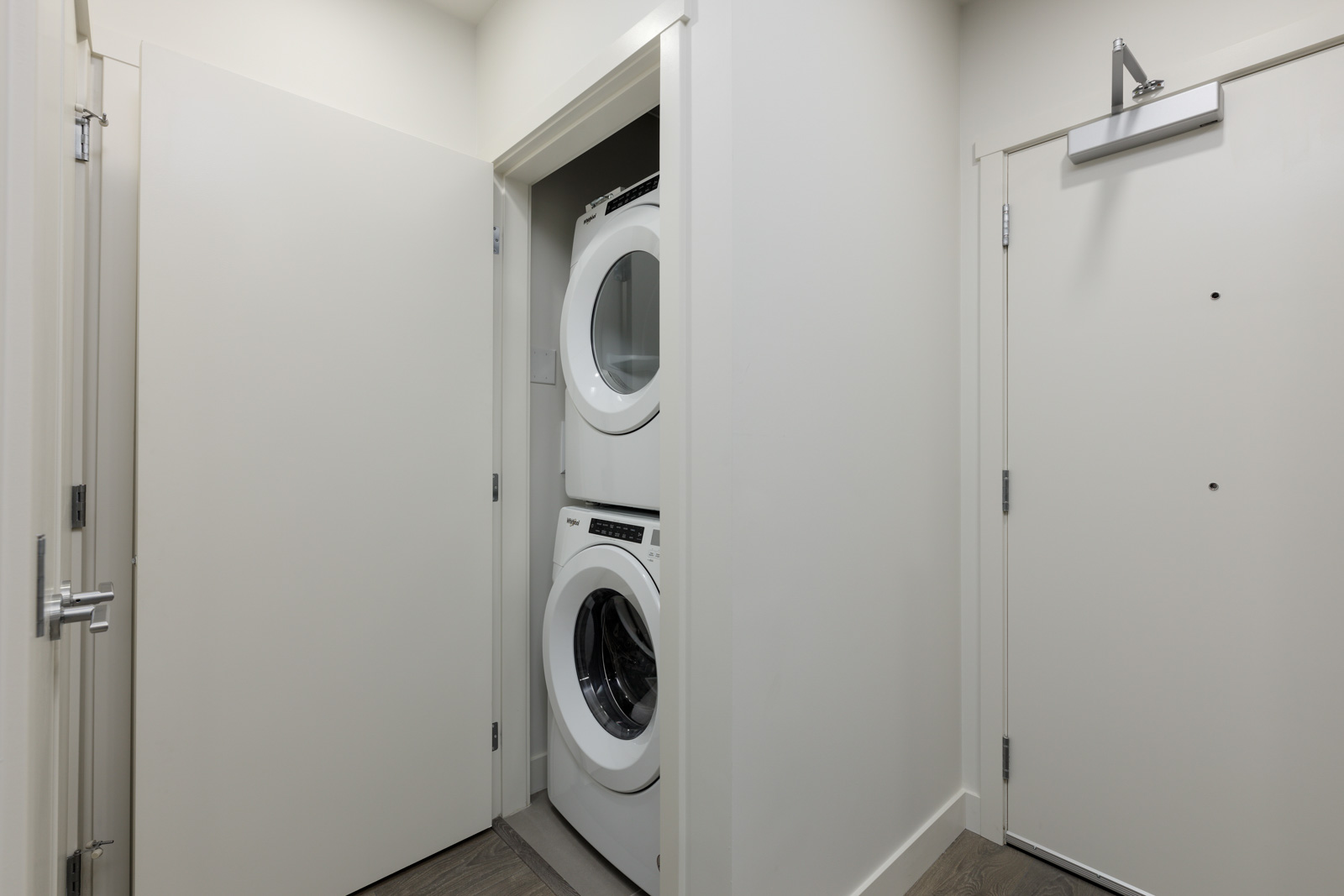 Stacked washer and dryer in a small closet next to a partially open door in a narrow hallway with white walls.