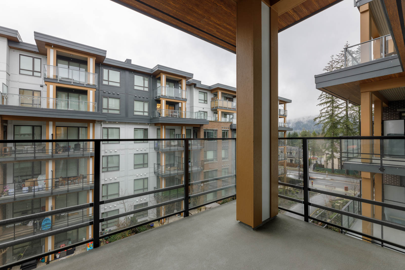 View from a balcony overlooking an apartment complex with modern architecture, featuring glass railings, multiple floors, and trees in the background. Cloudy sky overhead.