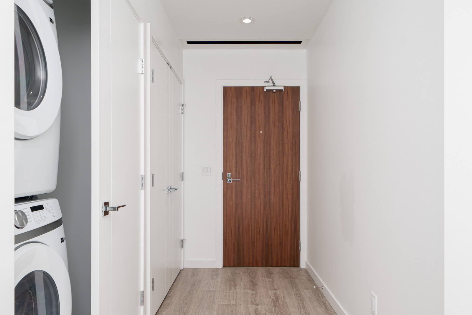 A hallway with a wooden door, white walls, and light wood flooring. A stacked washer and dryer are on the left side.