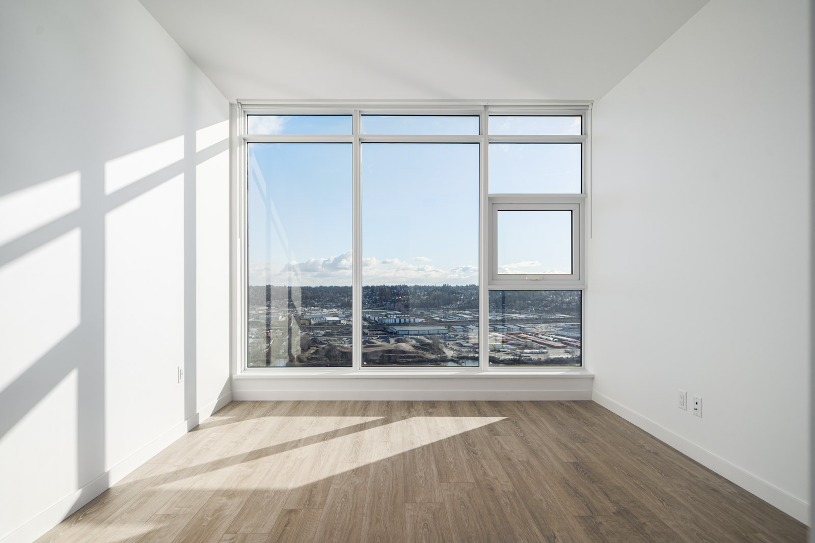 Empty room with large floor-to-ceiling windows, light wood flooring, and a view of a cityscape under a blue sky.