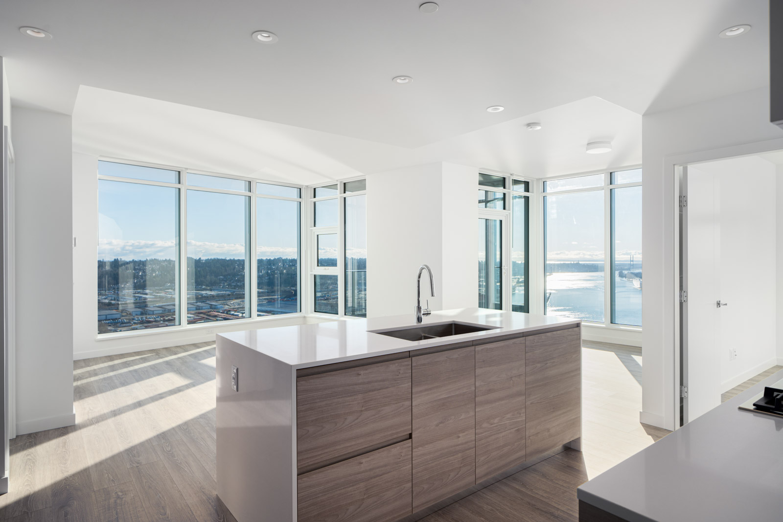 Modern apartment interior with large windows, ocean view, and an open-layout kitchen featuring a central island and wooden cabinets.