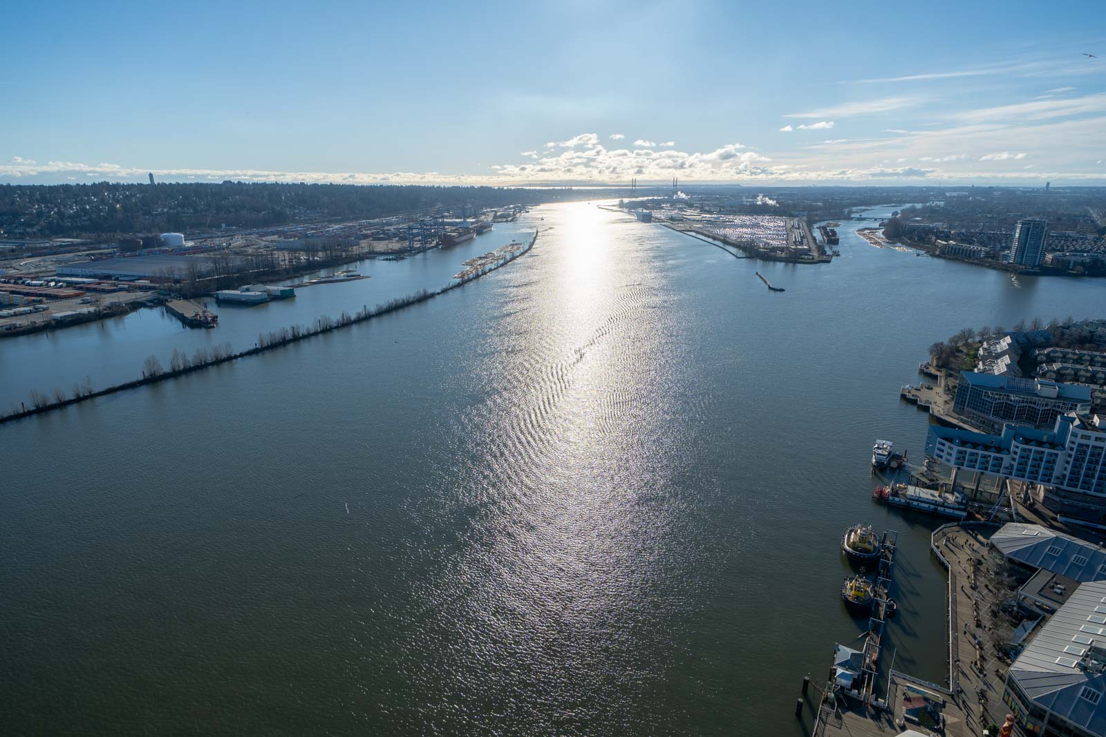 Aerial view of a wide river reflecting sunlight, surrounded by city buildings and docks, with a cloudy sky in the background.