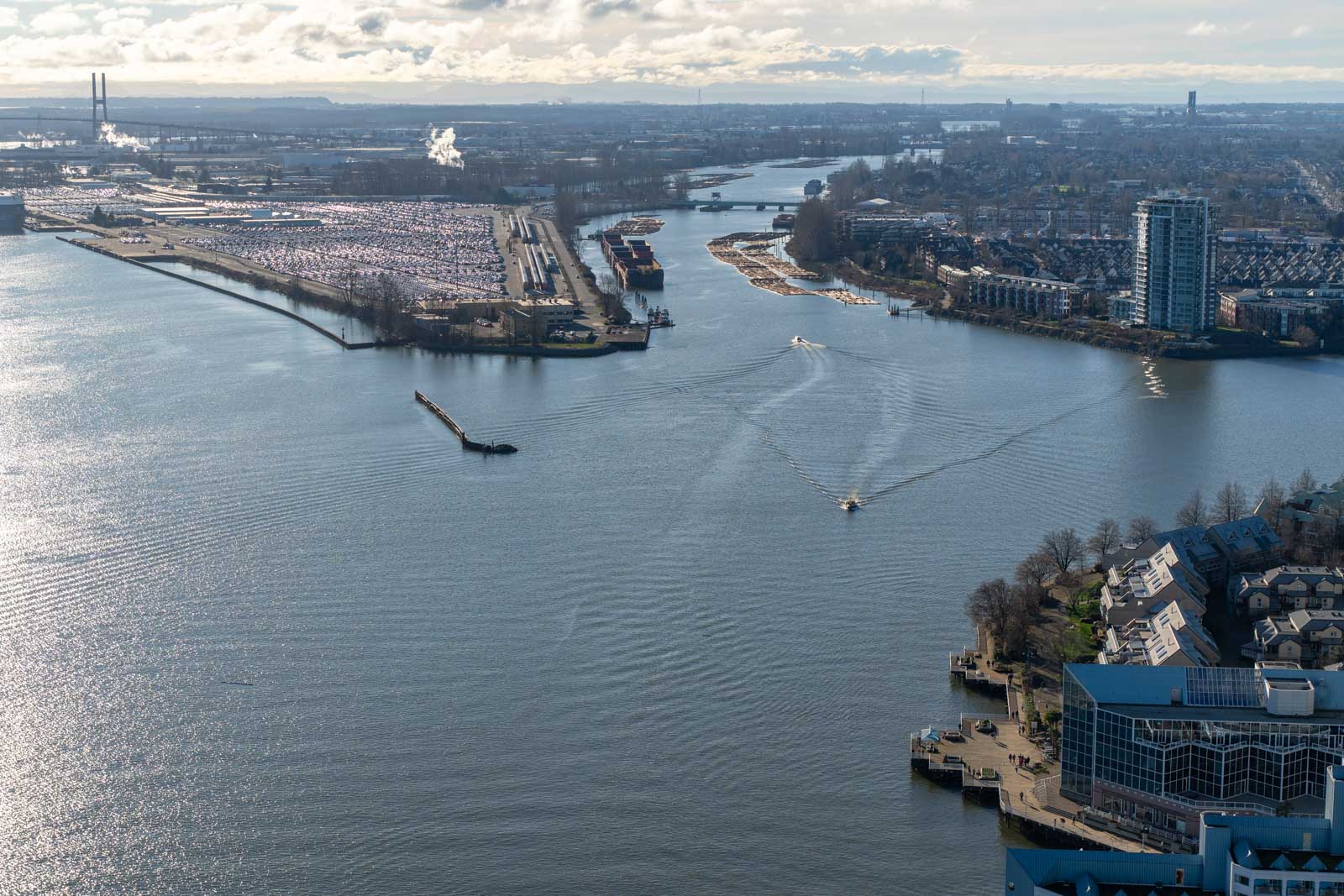 Aerial view of a river dividing a cityscape, with industrial areas on the left and residential buildings on the right. A boat creates ripples in the water.