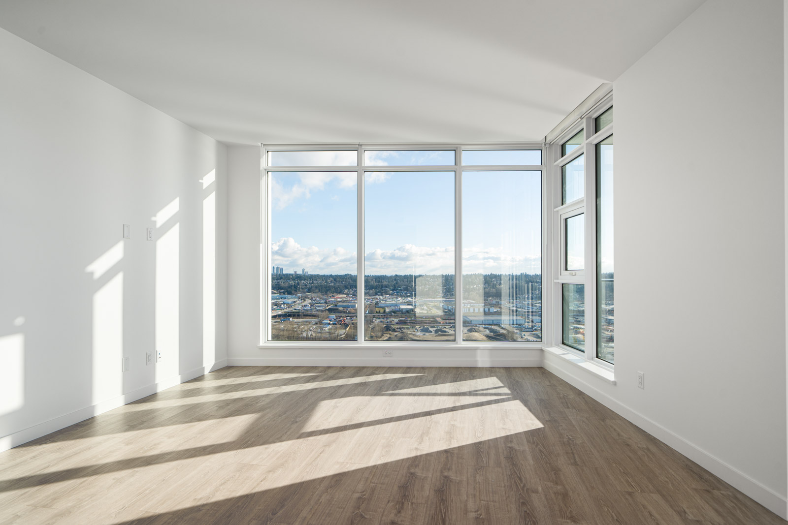 Empty room with large windows providing a view of a cityscape. Sunlight casts shadows on the wooden floor. Walls are painted white.