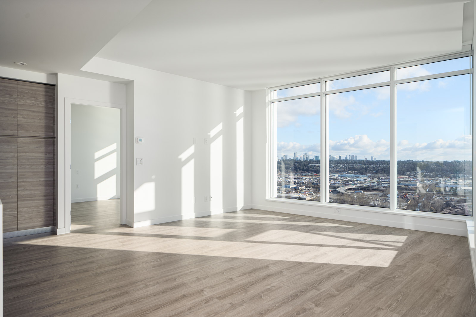 Bright empty room with large windows and wood flooring, offering a view of a cityscape under a blue sky.