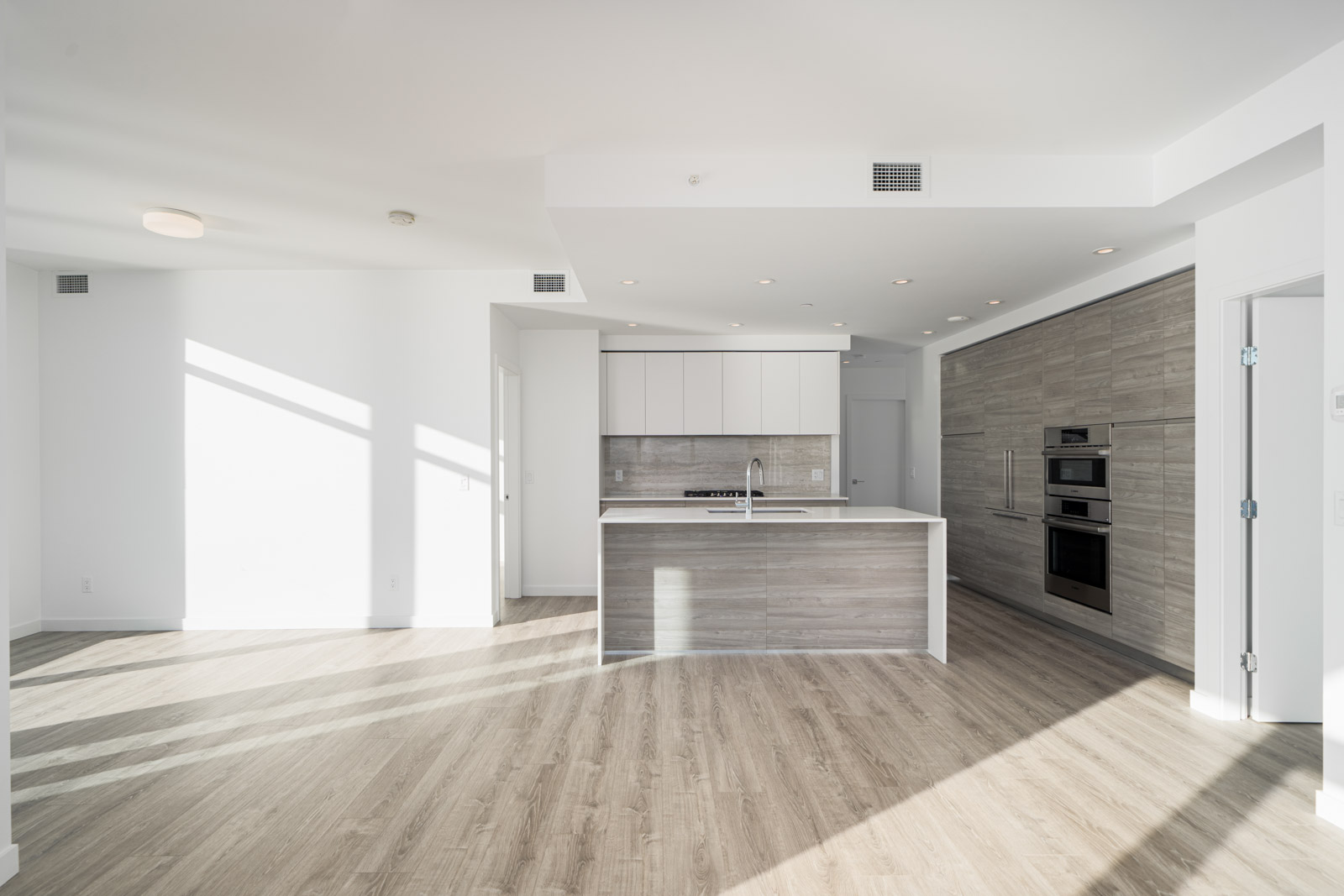 Modern kitchen with light wood flooring, white cabinets, and built-in oven. Central island with sink and minimalist design. Bright natural light from large windows.