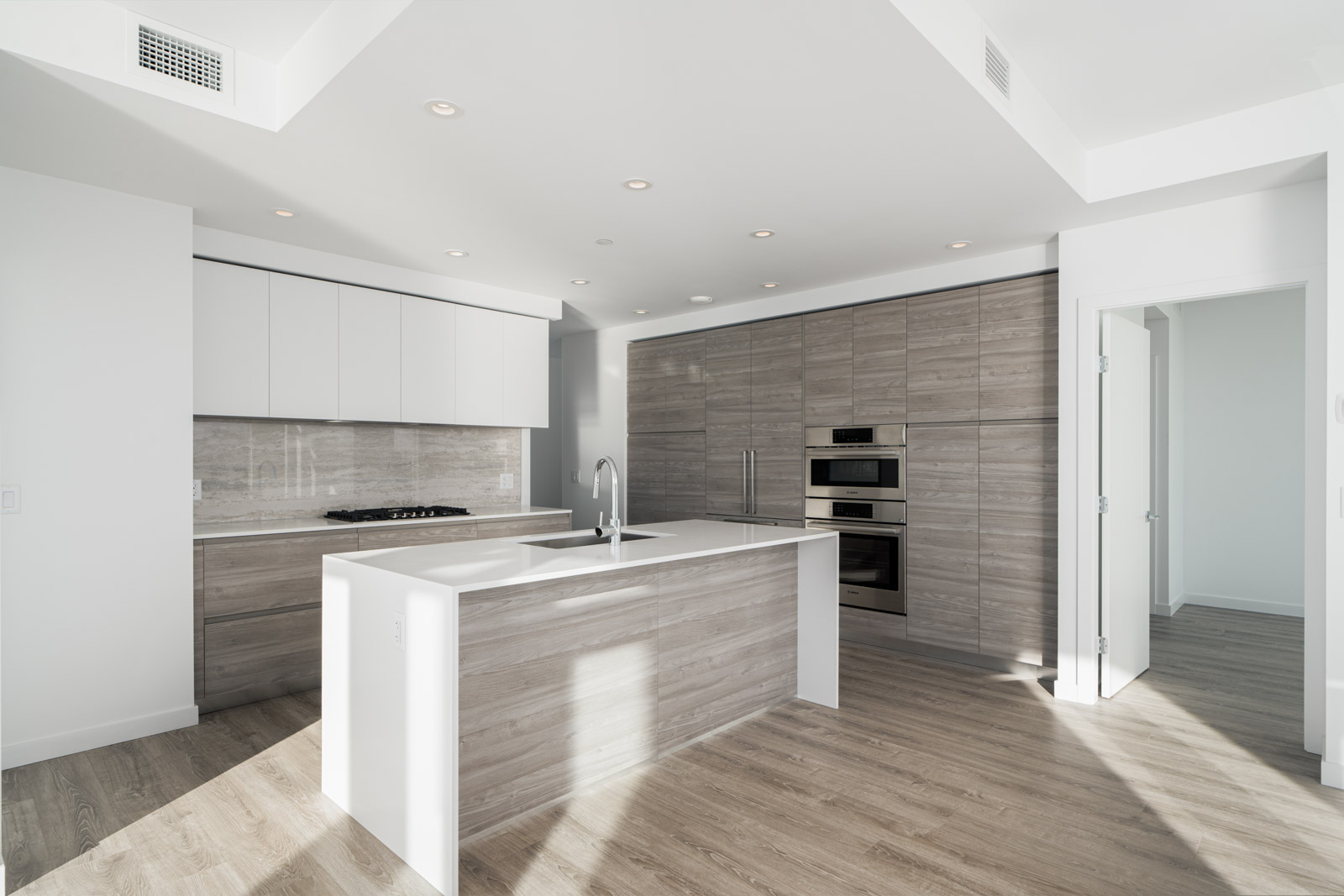 Modern kitchen with a central island, built-in oven, light wood cabinets, and recessed lighting. White walls and ceiling, with an open doorway on the right.