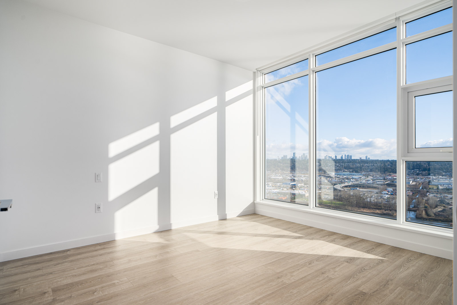 Bright empty room with large windows, wood flooring, and a view of a cityscape under a clear sky.