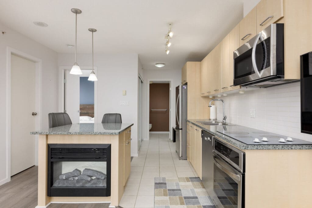 Modern kitchen with light wood cabinets, stainless steel appliances, a glass-top stove, a double sink, and a wall-mounted microwave. Two pendant lights hang over a peninsula with two stools.