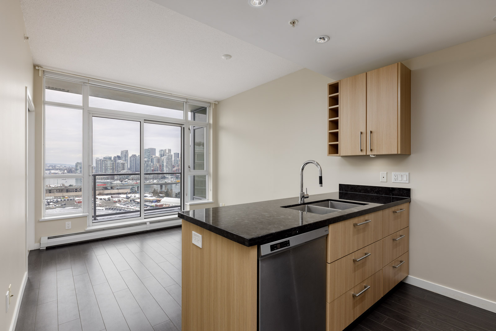 Modern kitchen with wooden cabinets, black countertop, and stainless steel sink. Large windows offer a cityscape view in the background.