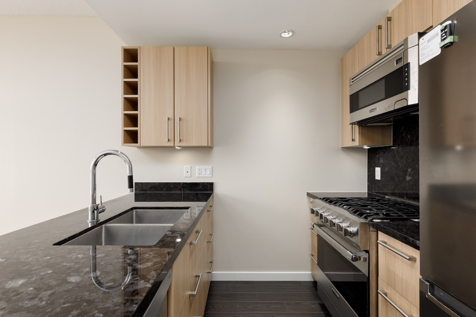 Modern kitchen with light wood cabinets, black countertops, stainless steel appliances, and a built-in wine rack. Includes a sink on a central island, and a gas stove and microwave on the right.