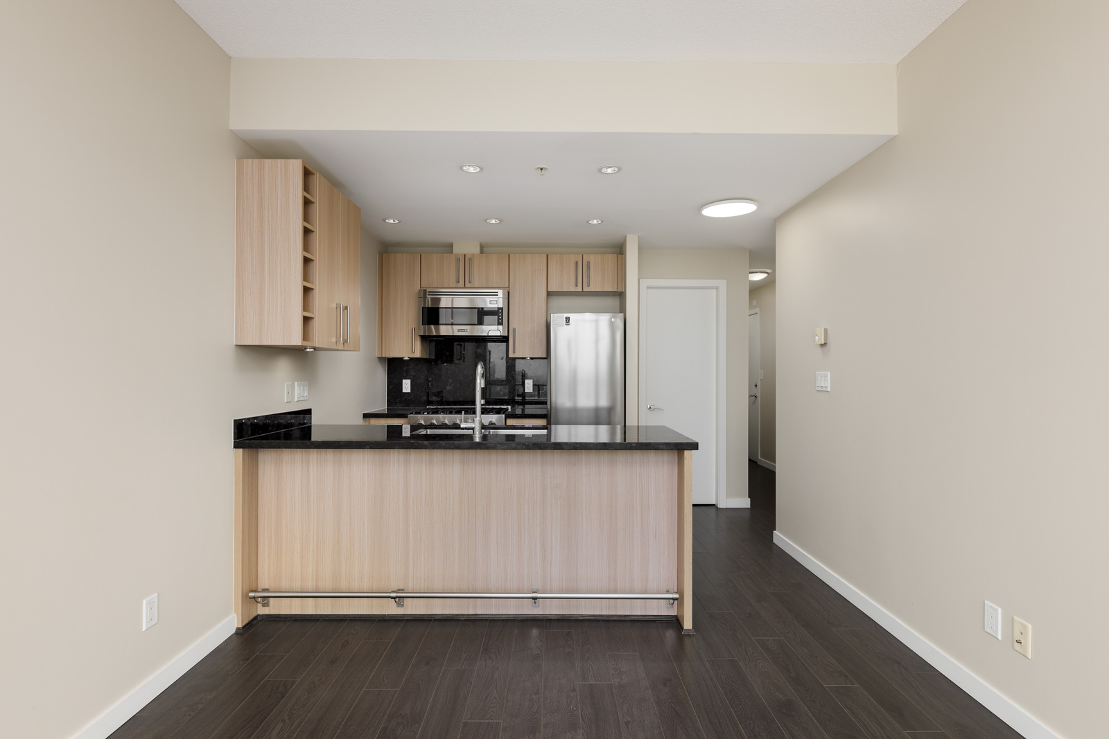 Modern kitchen with light wood cabinetry, black granite countertops, stainless steel appliances, and a small bar counter. Dark wood flooring and neutral wall paint complete the space.