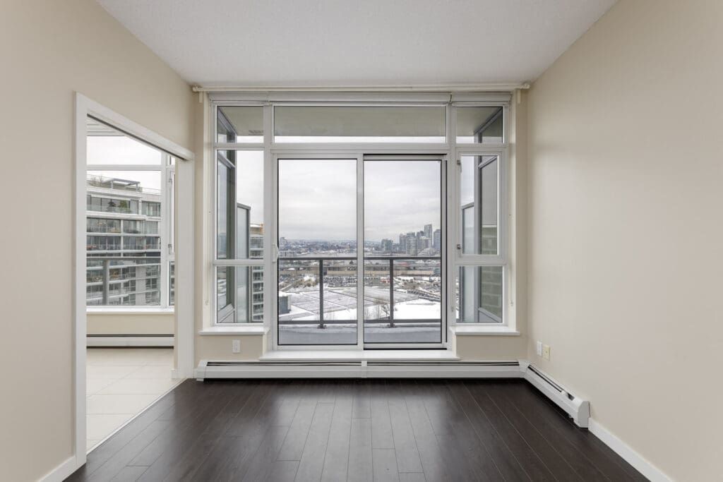 Empty room with dark flooring, large windows, and a view of the city. Adjacent room with white flooring visible through an open doorway.