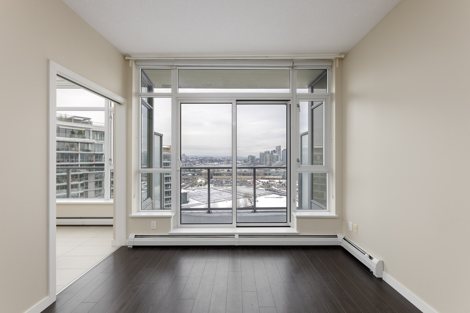 Empty room with dark flooring, large windows, and a view of the city. Adjacent room with white flooring visible through an open doorway.