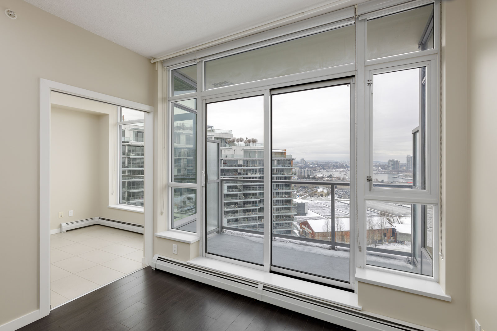 Bright room with large windows and sliding glass doors leading to a balcony. Overcast sky and cityscape visible outside. Light-colored walls and dark wooden floors inside.
