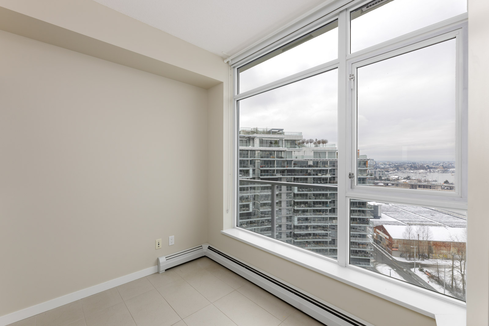 Empty room with beige walls and large window showing a cityscape with tall buildings and a river in the background.