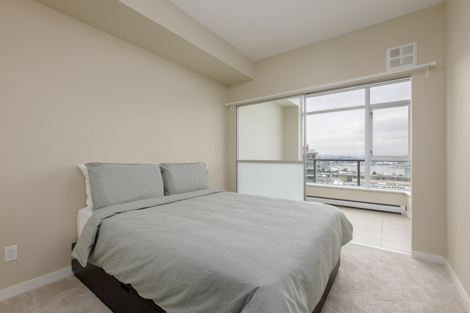 Modern bedroom with a neatly made bed and a large window offering a cityscape view. Walls are painted beige, and there is natural light entering the room.