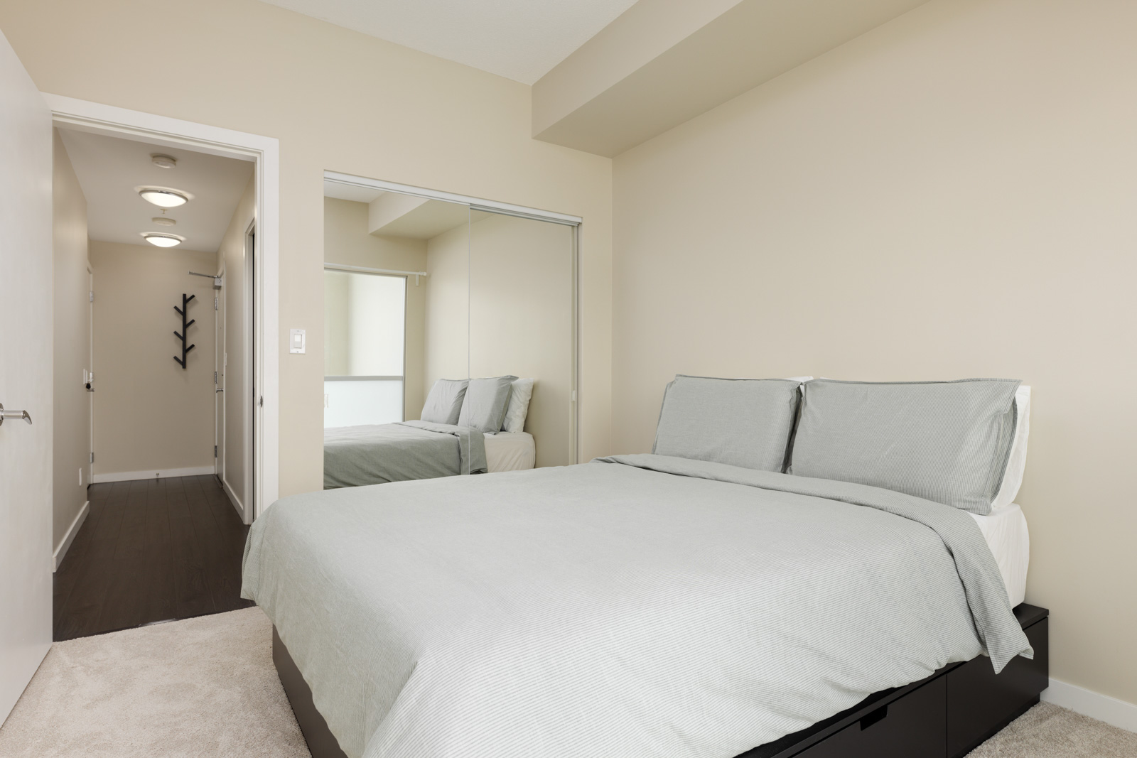 A bedroom with a neatly made bed, gray bedding, a mirrored closet, beige walls, and a view of a hallway with two ceiling lights and a black wall hook.