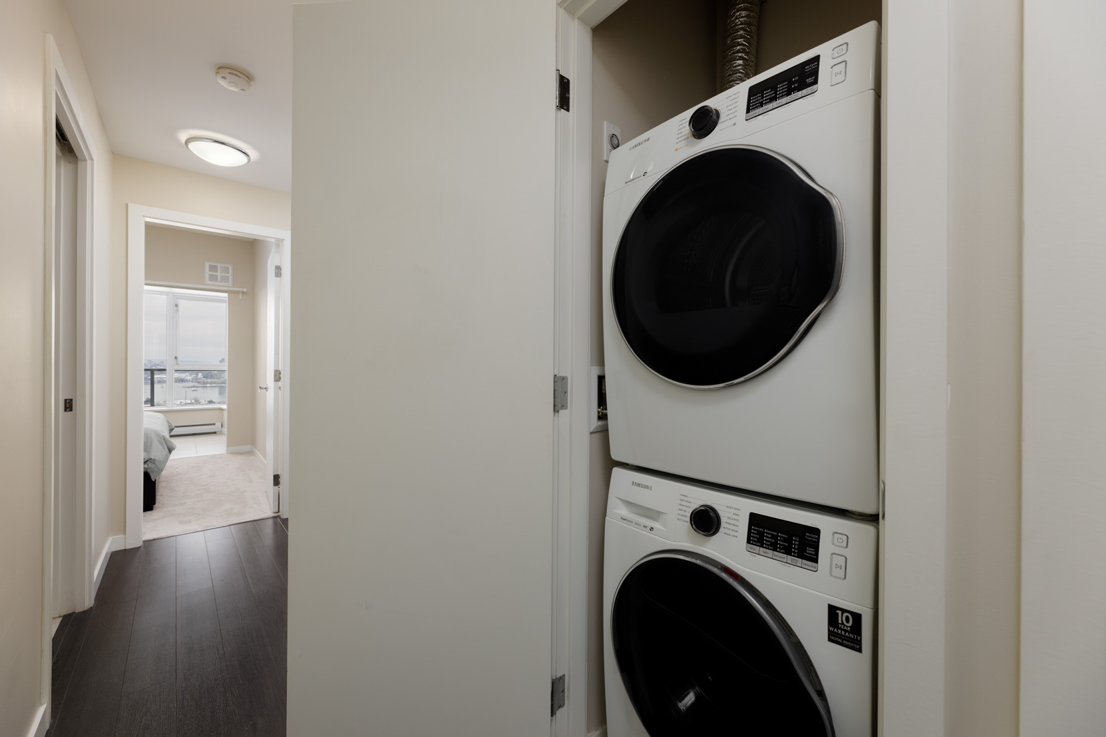 Stacked washer and dryer in a small laundry closet, with a hallway leading to a bedroom visible to the left.