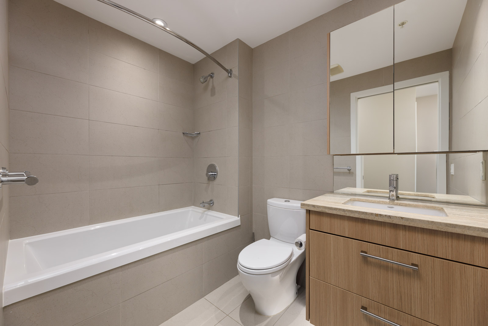 Modern bathroom with beige tiles, a bathtub with shower fixtures, a toilet, and a wooden vanity with a mirror cabinet above the sink.
