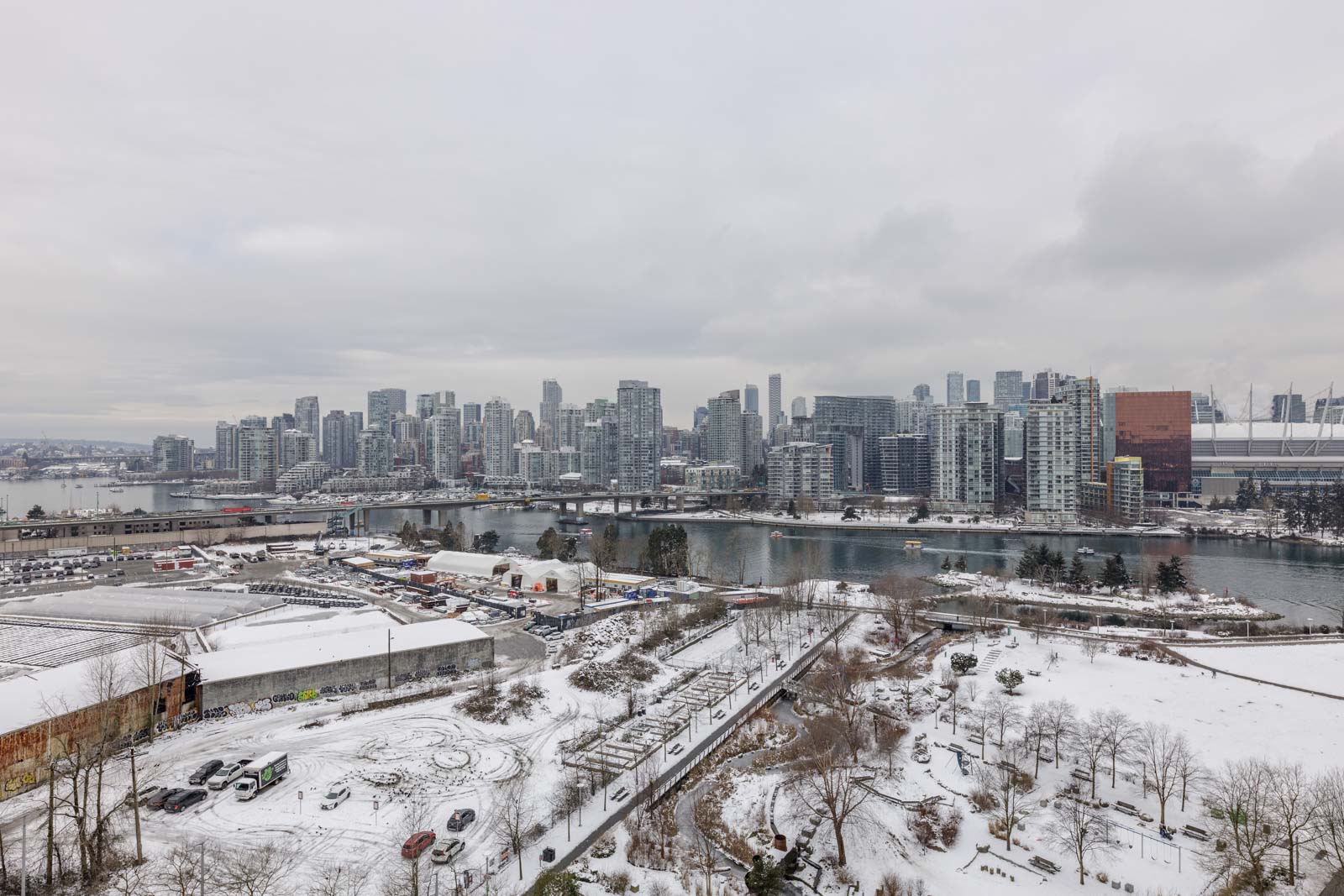 Snow-covered cityscape with a river, modern buildings, and overcast sky. Urban areas and waterfront visible under a light layer of snow.