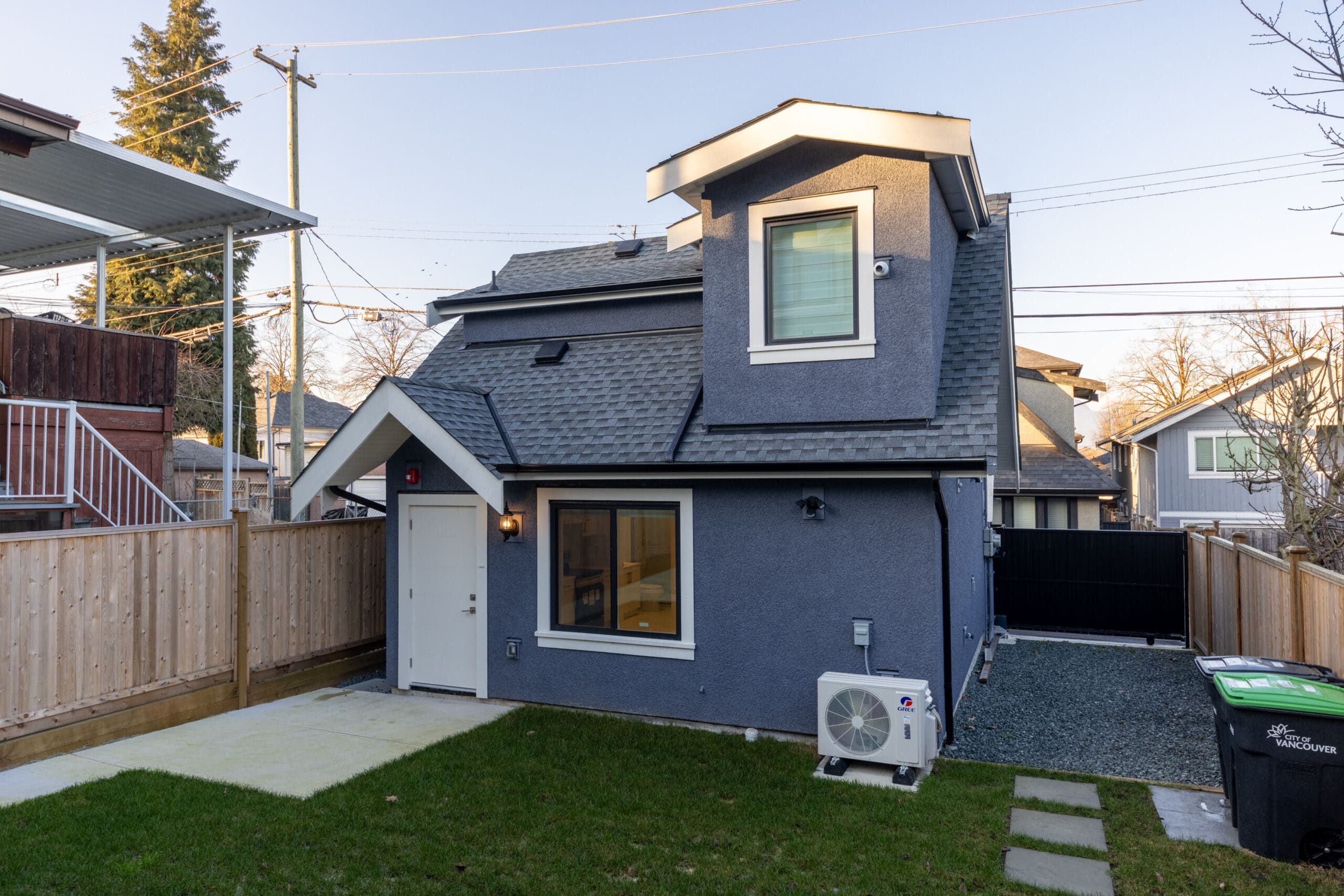 A small, two-story gray house with a fenced yard, green grass, and gravel area. An air conditioning unit is mounted on the side, and a trash bin is visible near the driveway.