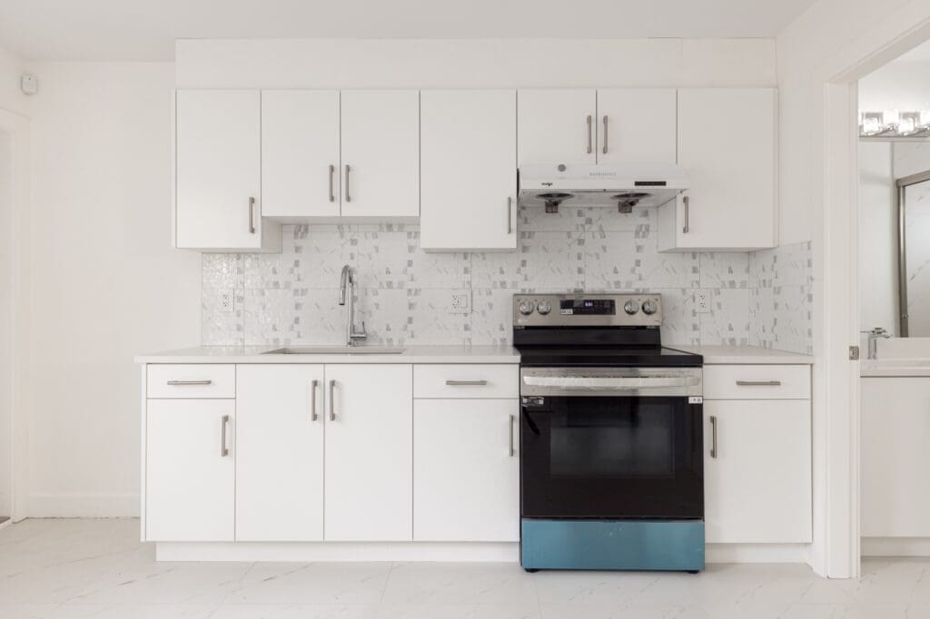 Modern kitchen with white cabinets, silver handles, tiled backsplash, stainless steel stove and range hood, and a single sink.