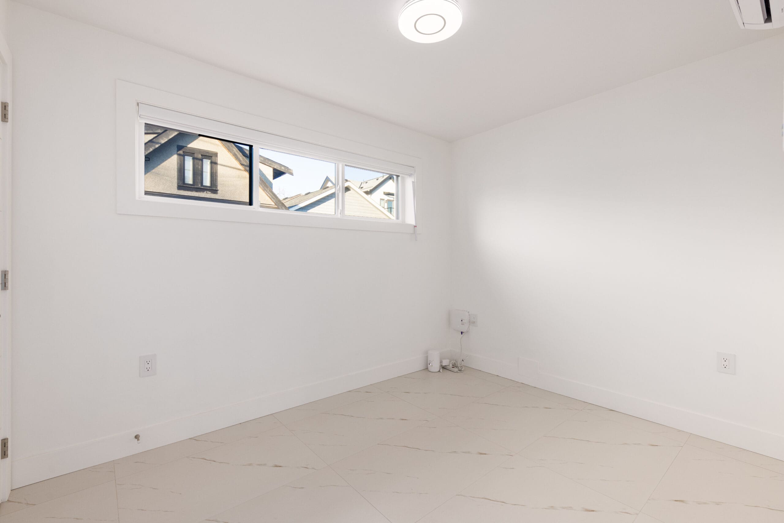 Empty white room with tiled floor, a small horizontal window showing rooftops outside, and minimal light fixture on the ceiling.