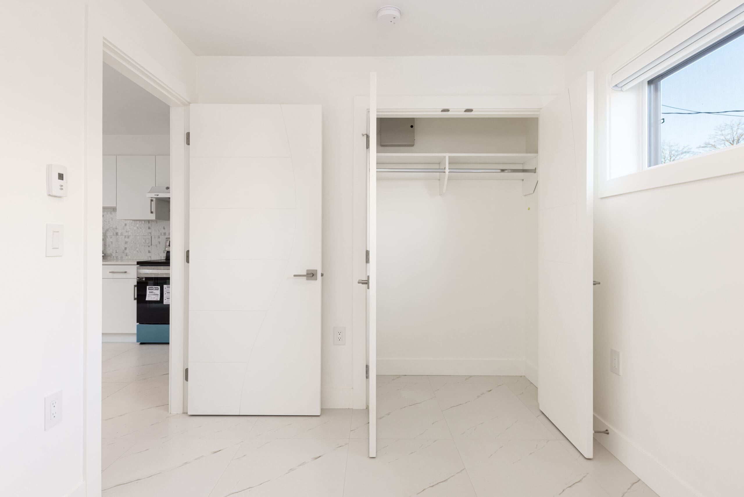 Empty white room with open closet doors, tiled floor, and a view of a kitchen through an open door.