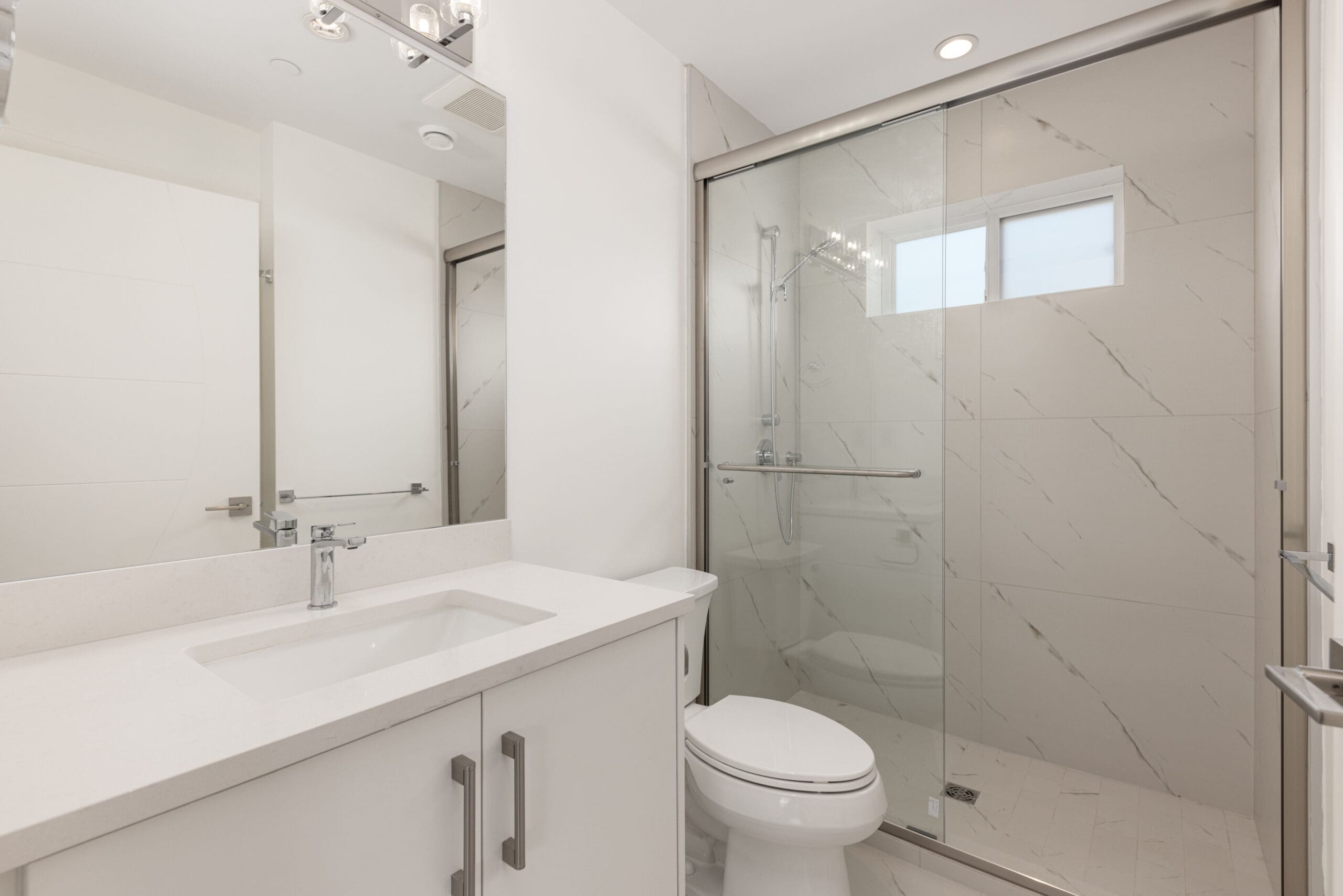 Modern bathroom with a white vanity, sink, toilet, and a glass-enclosed shower. Light-colored tiles and walls create a clean, spacious look.