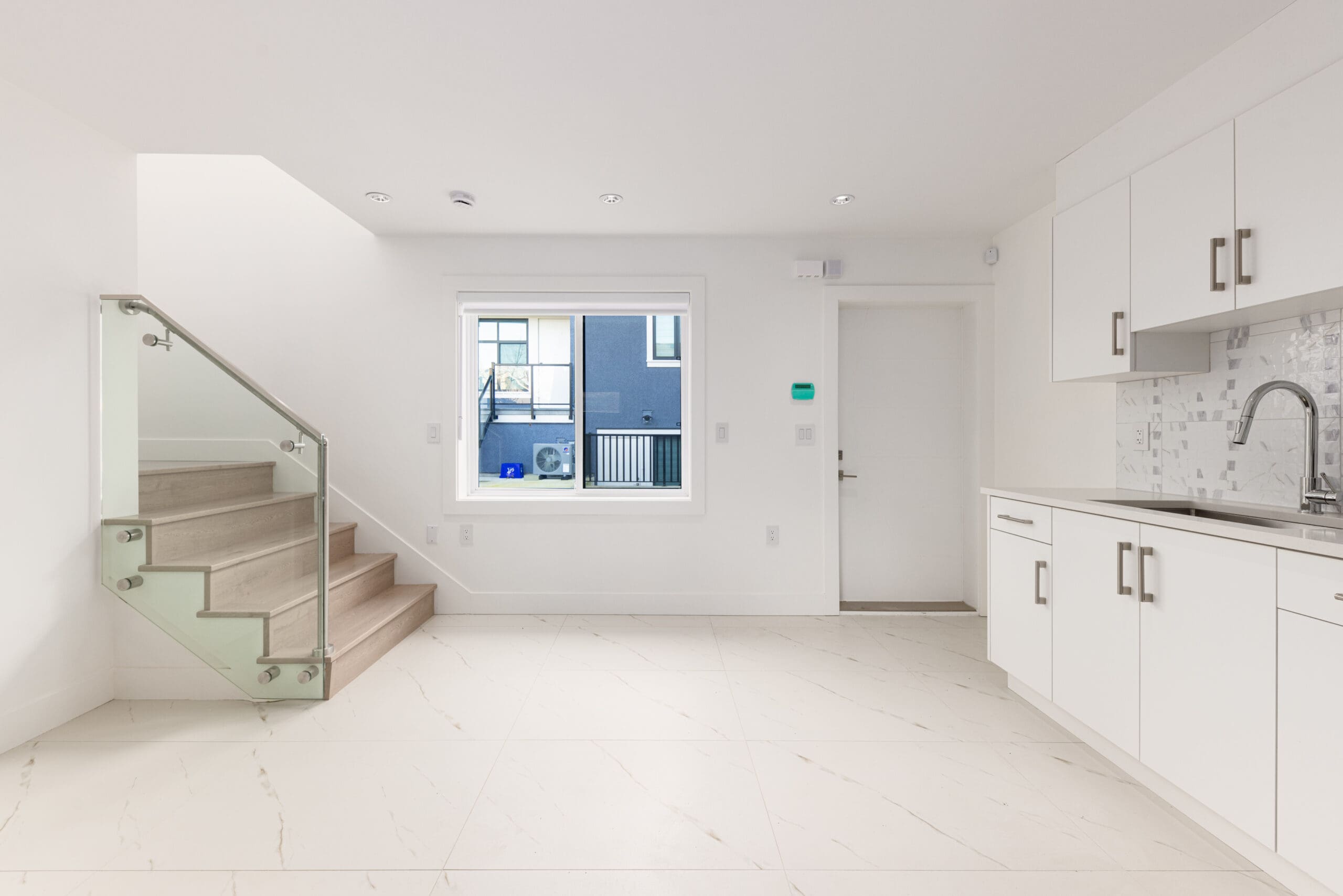 Modern, minimalist kitchen with white cabinets, marble floor, sink, backsplash, and a staircase leading upward. Large window and white walls enhance natural light.