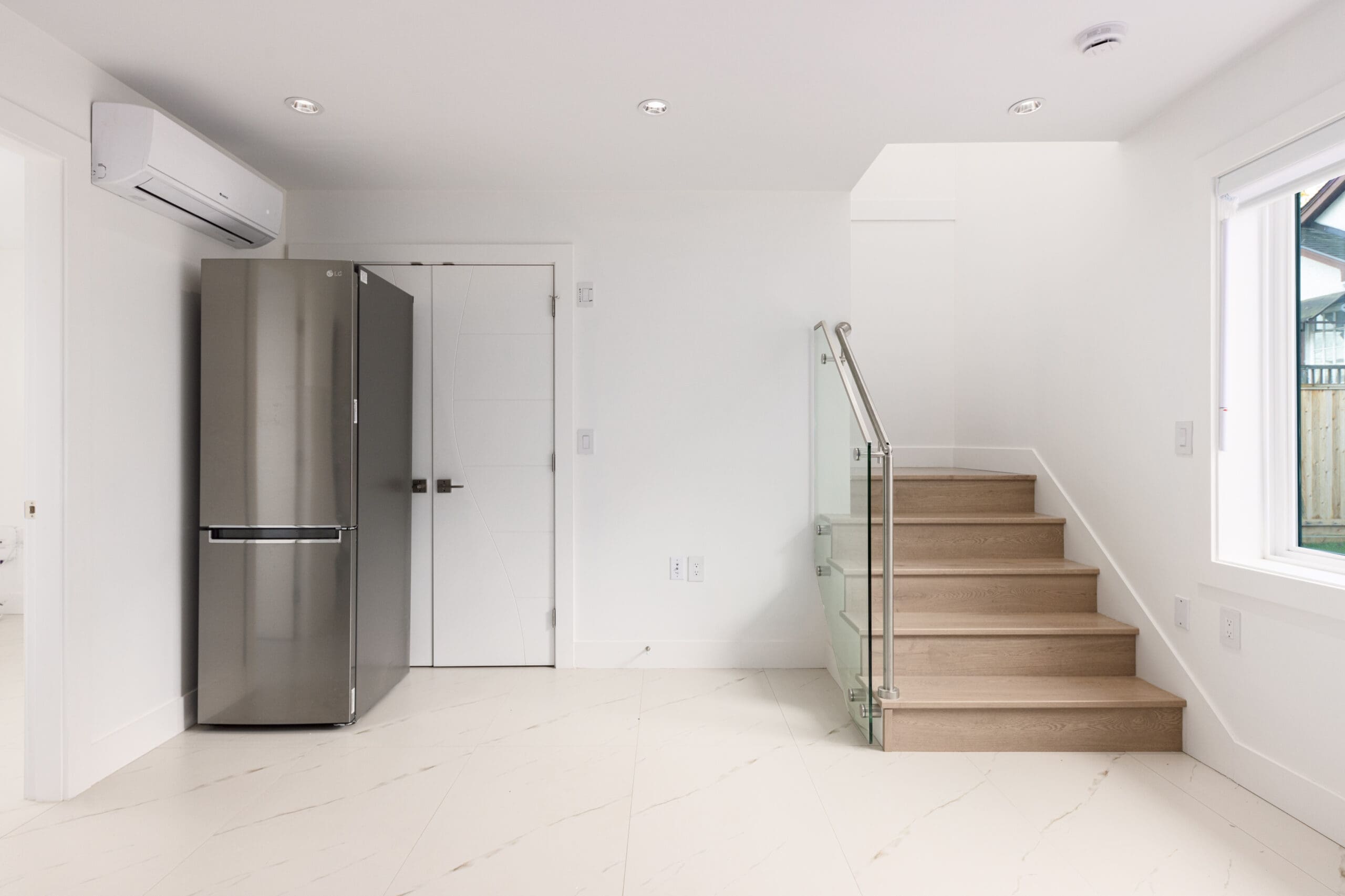 Minimal kitchen with stainless steel refrigerator, white walls, wooden stairs with glass railing, and tile flooring. Air conditioning unit above fridge and window on the right.