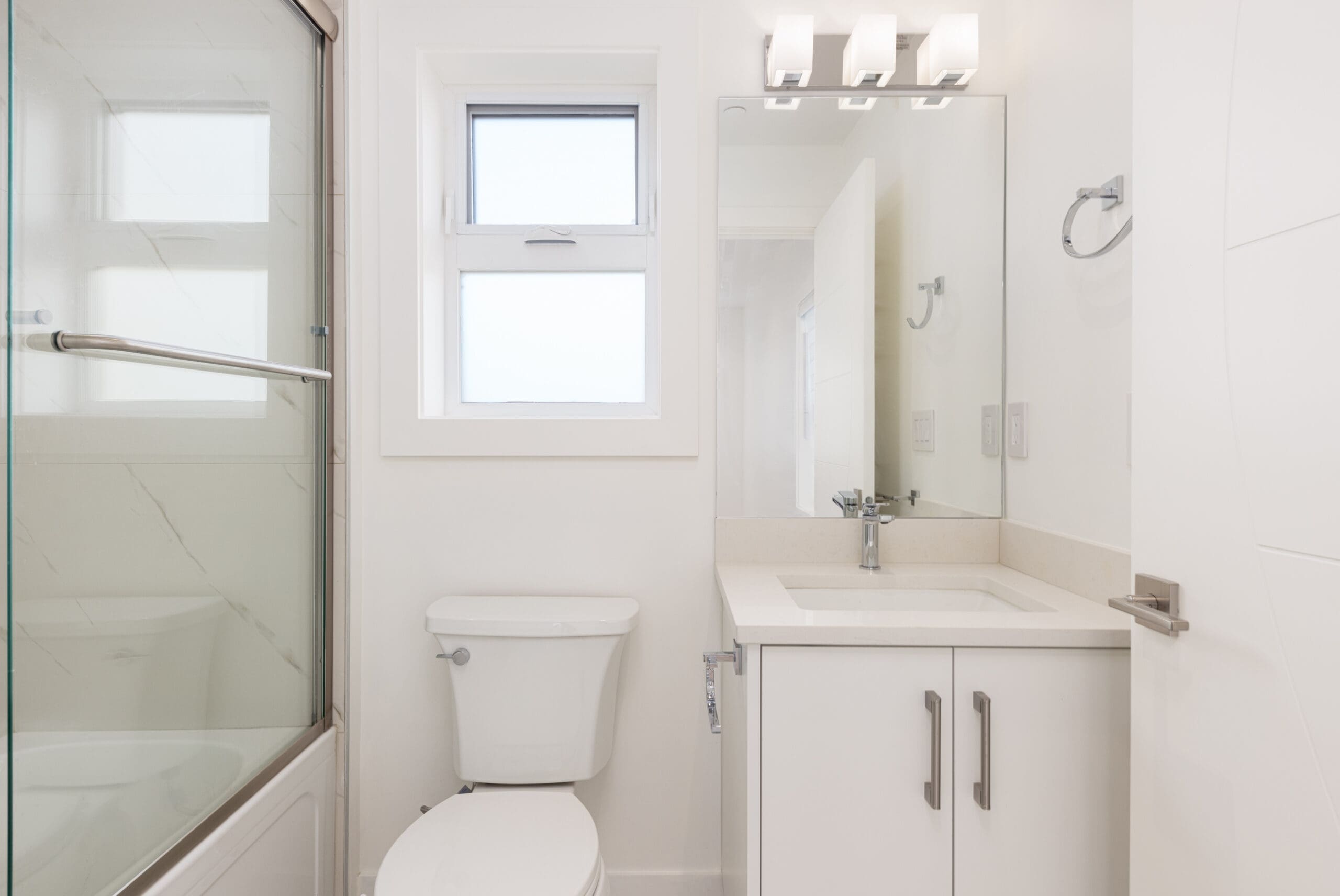 A modern bathroom with a white sink and cabinet, a mirror, a toilet, and a shower with glass doors. The room is lit by a three-bulb light fixture above the mirror.