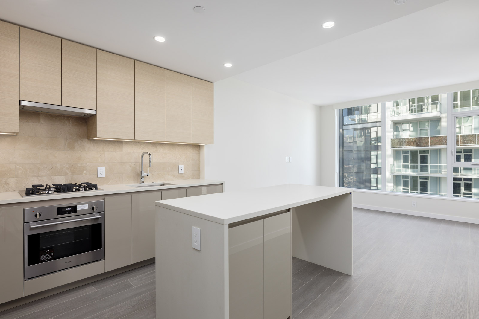 Modern kitchen with light wood cabinets, beige countertops, stainless steel appliances, and an island. Adjacent is a living area with large windows and light wood flooring.