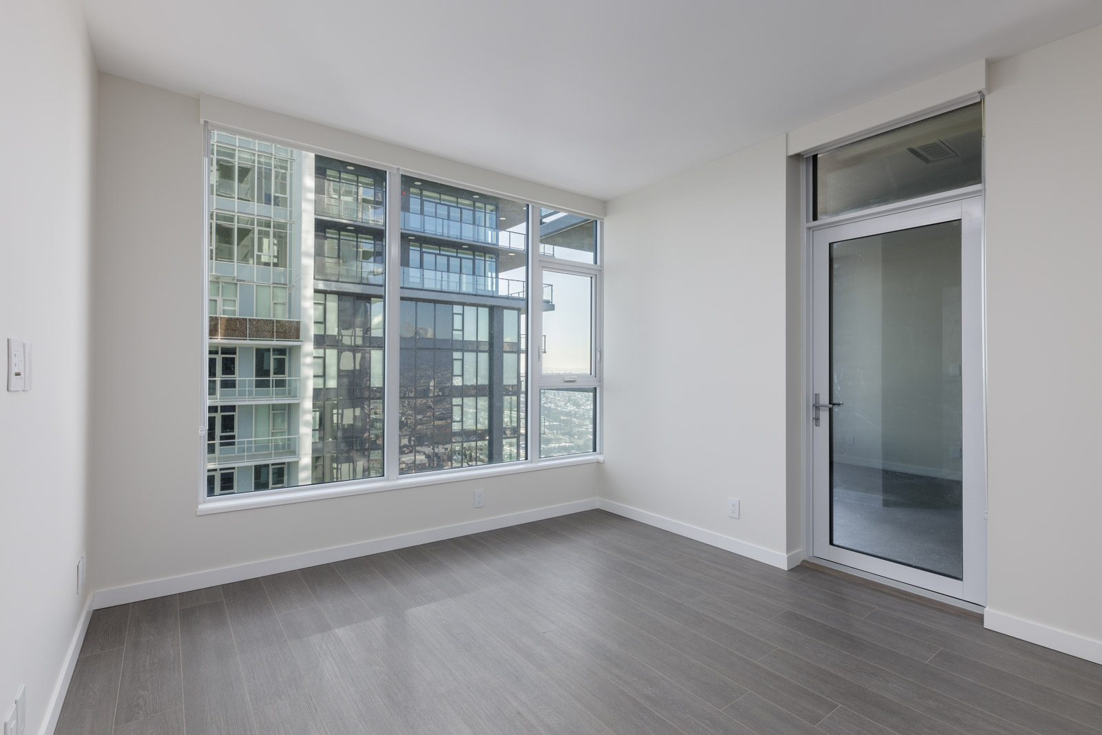 Empty, modern room with large windows and a glass door, featuring light gray wood flooring and white walls, overlooking a cityscape from a high-rise building.