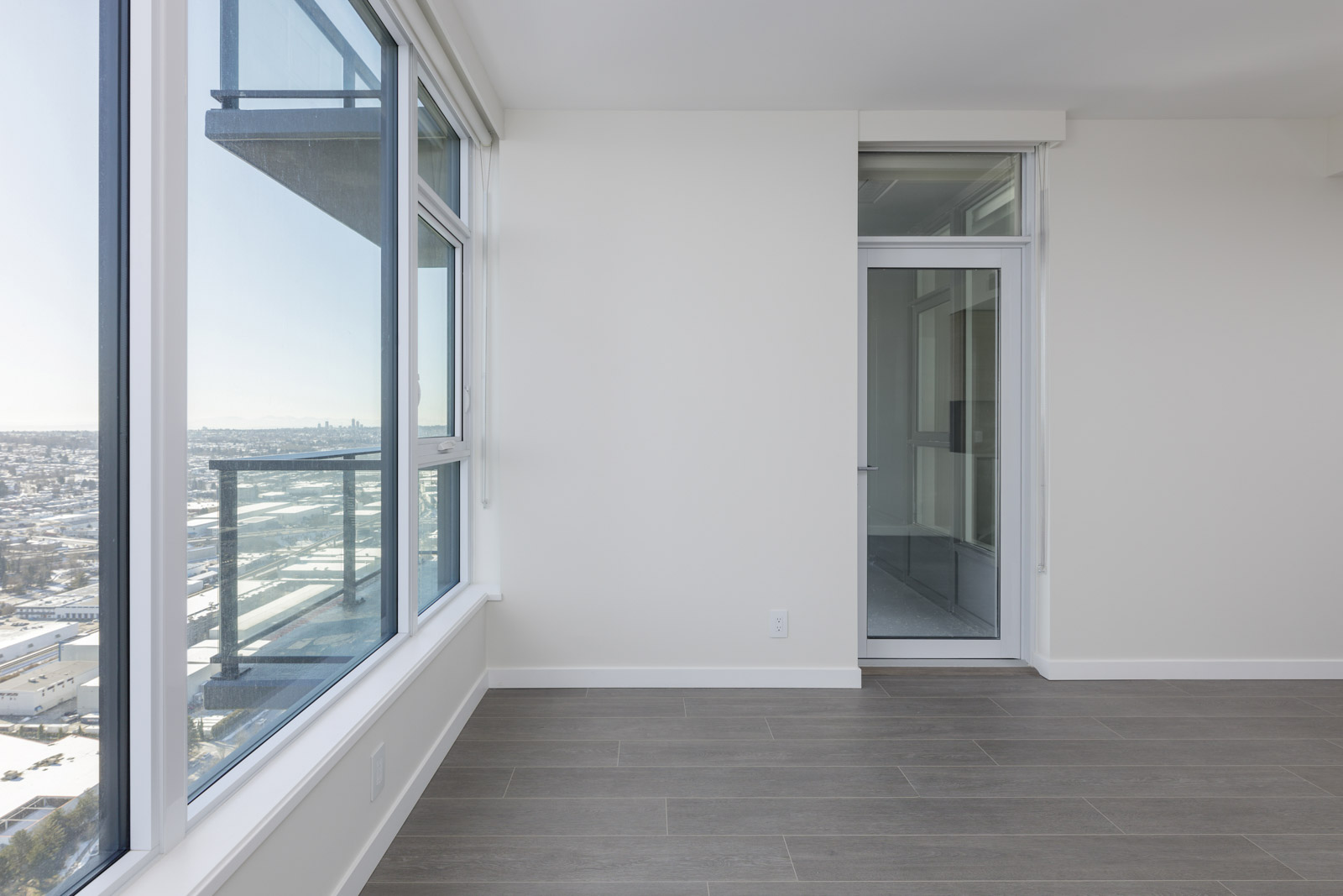 Bright, empty room with large windows and a glass door leading to a balcony. The floor has wood-like tiles, and the walls are white. Cityscape visible through the windows.