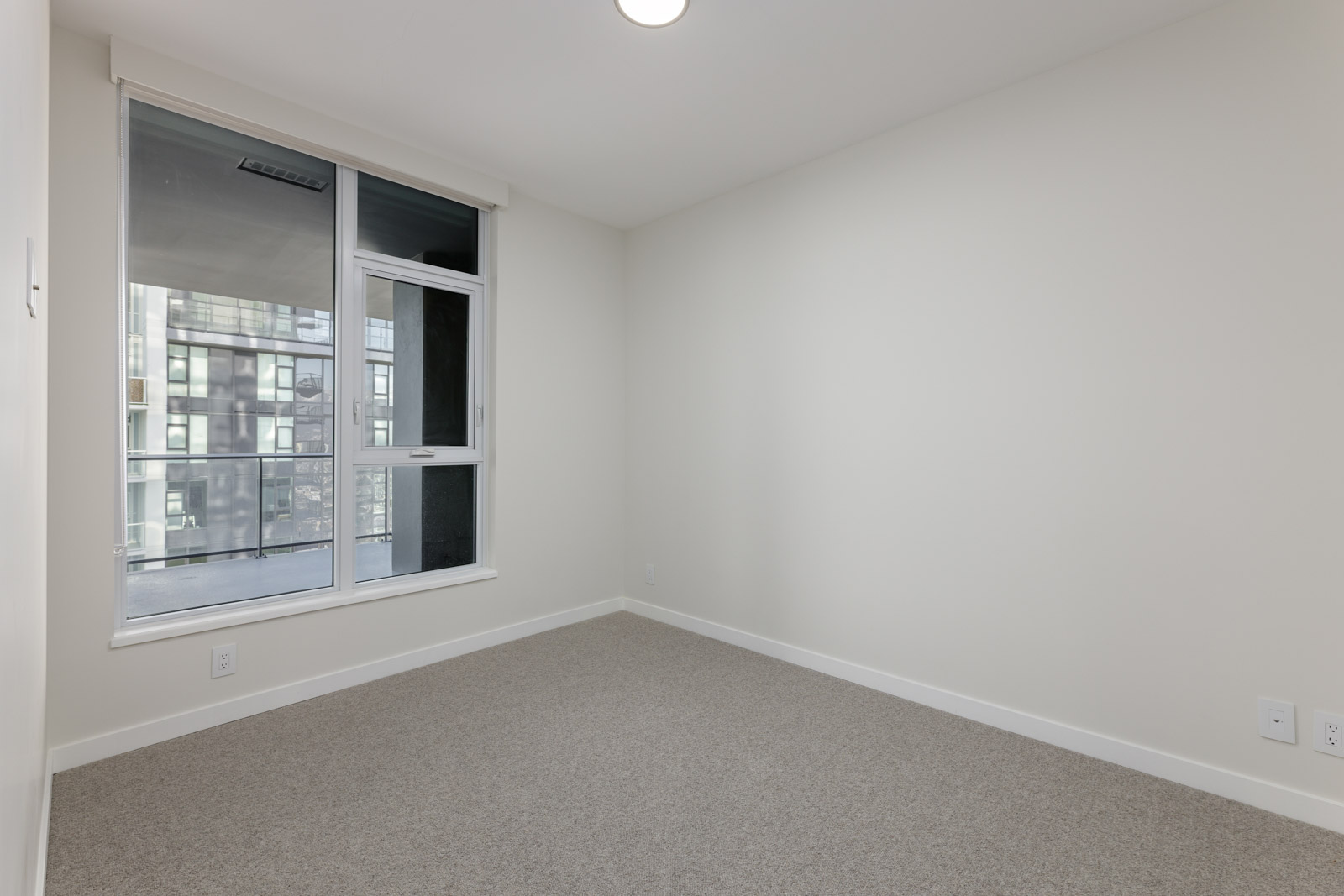 Empty room with beige carpet, white walls, and a large window showing an exterior urban view.