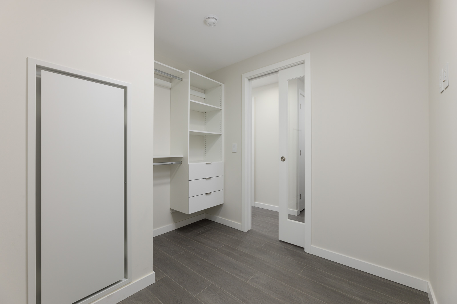 Empty room with white walls, light wood flooring, a built-in closet with shelves and drawers, and sliding doors.