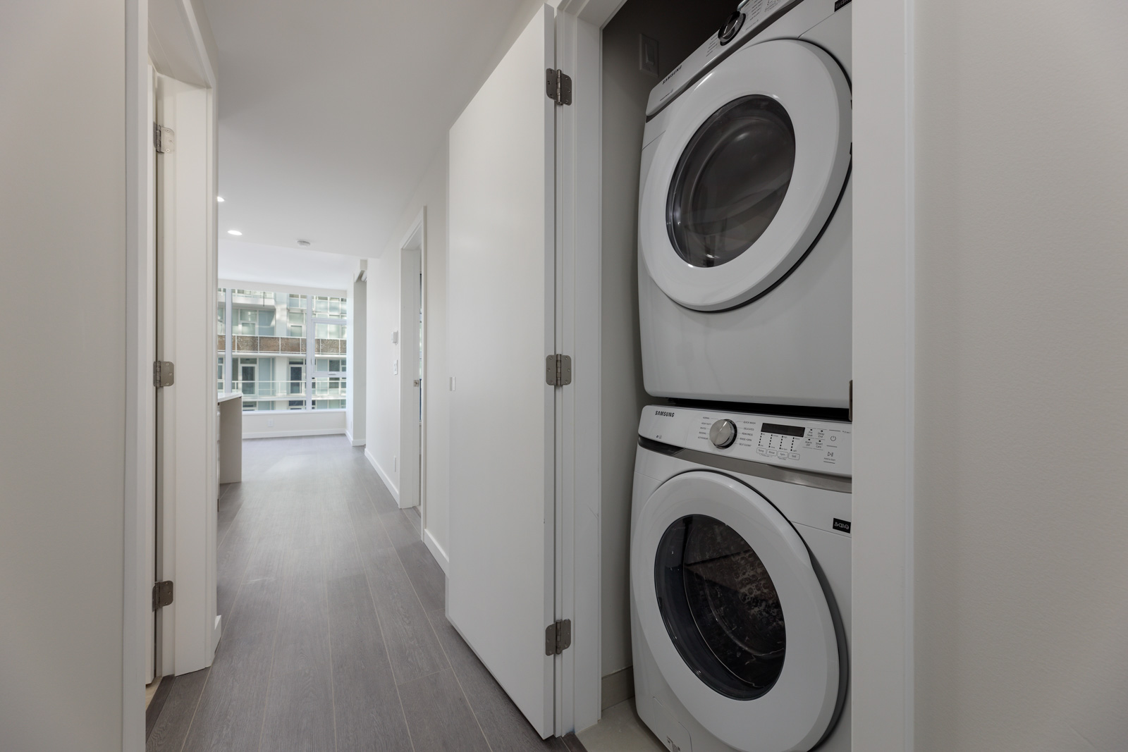Stacked washer and dryer in a narrow laundry closet beside a corridor leading to a room with large windows.