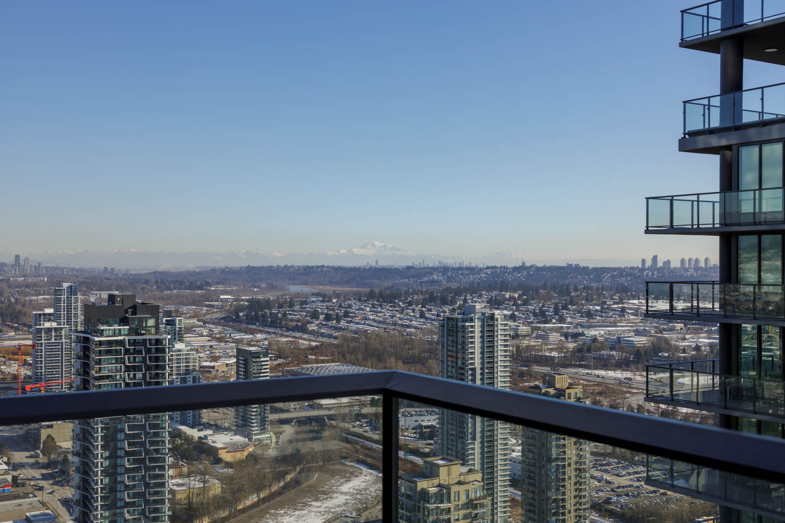 Cityscape view from a balcony with high-rise buildings in the foreground and mountains in the distant background under a clear blue sky.