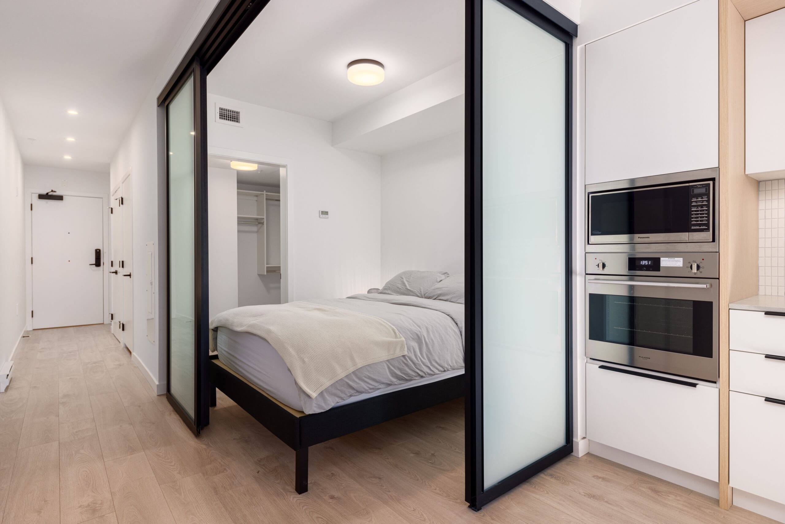 Compact bedroom with a bed and nightstand, separated by sliding glass doors from a kitchenette area with built-in appliances, including a microwave and oven.
