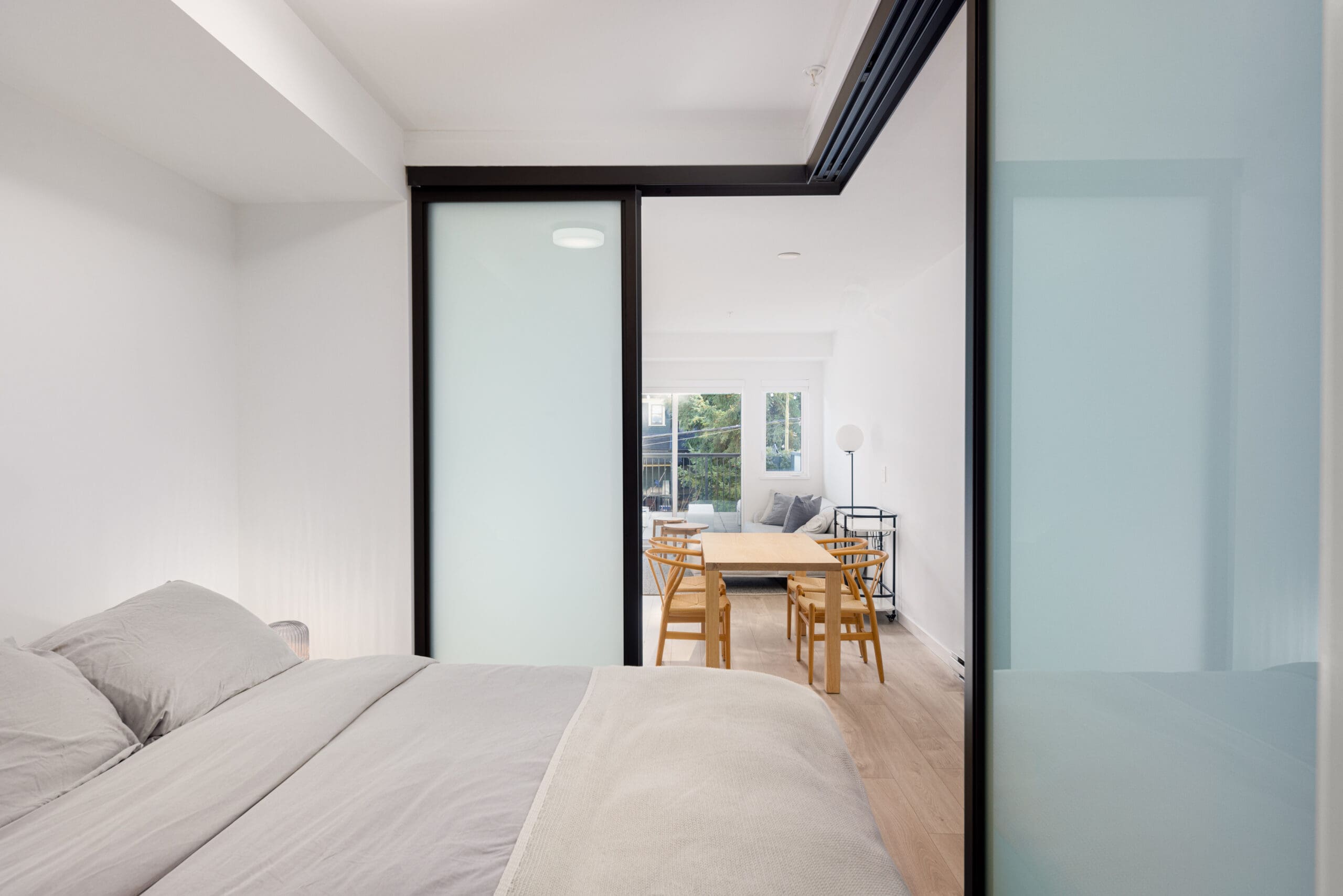 View from a bedroom through sliding frosted glass doors into a minimalist living and dining area with a wooden table, chairs, and a sofa, showcasing a bright, airy interior design.