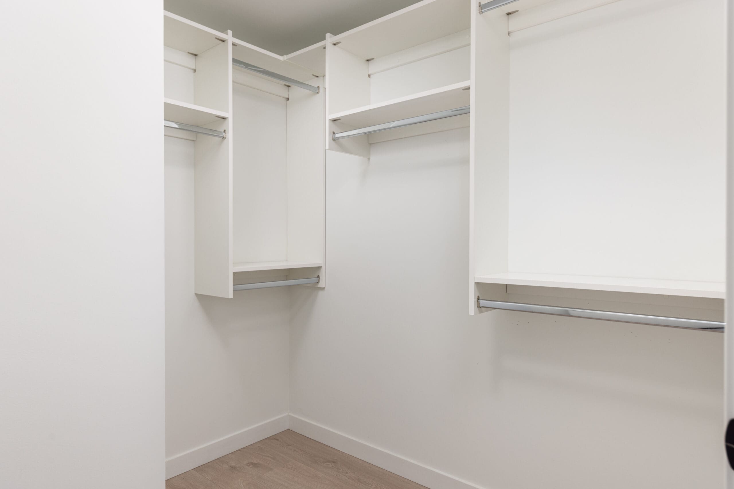 Empty walk-in closet with white shelves and hanging rods, light wooden floor, and white walls.