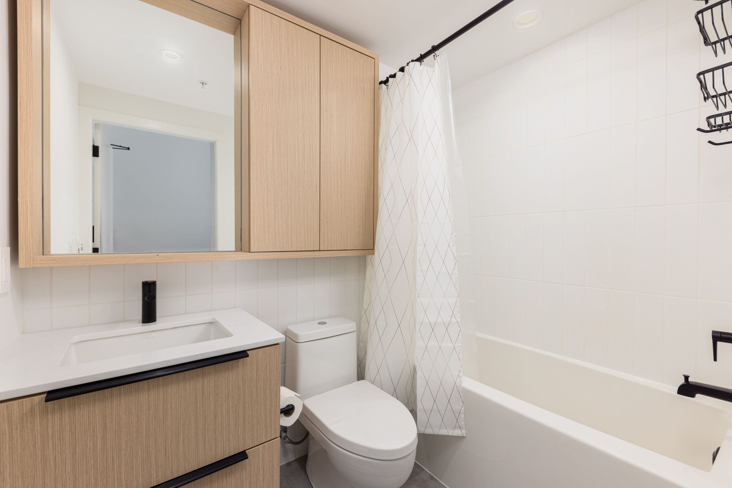 Modern bathroom with light wood cabinets, white sink, toilet, and bathtub with white shower curtain. Black fixtures and a large mirror are present.