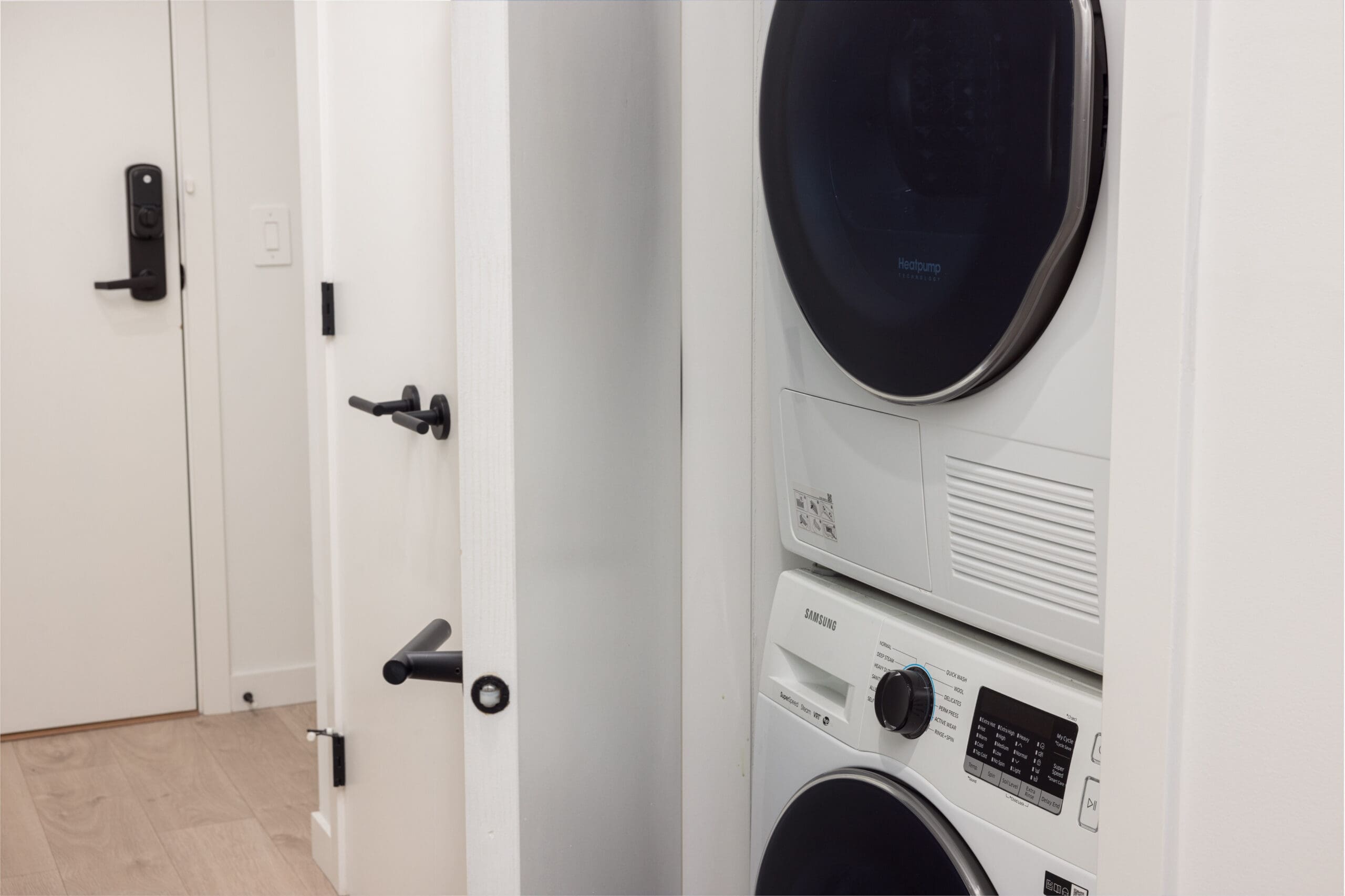 Stacked washer and dryer unit in a narrow laundry space with white walls and light wooden floor. A partially open door with black handles is visible on the left.