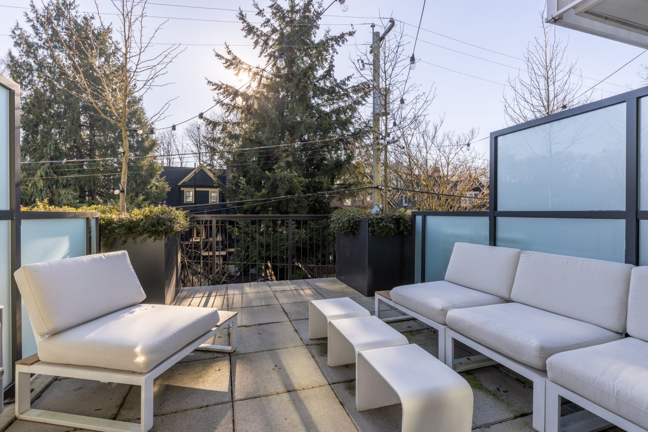 Outdoor patio with white cushioned seating, small tables, and frosted glass panels. Trees and a house are visible in the background, under clear skies.