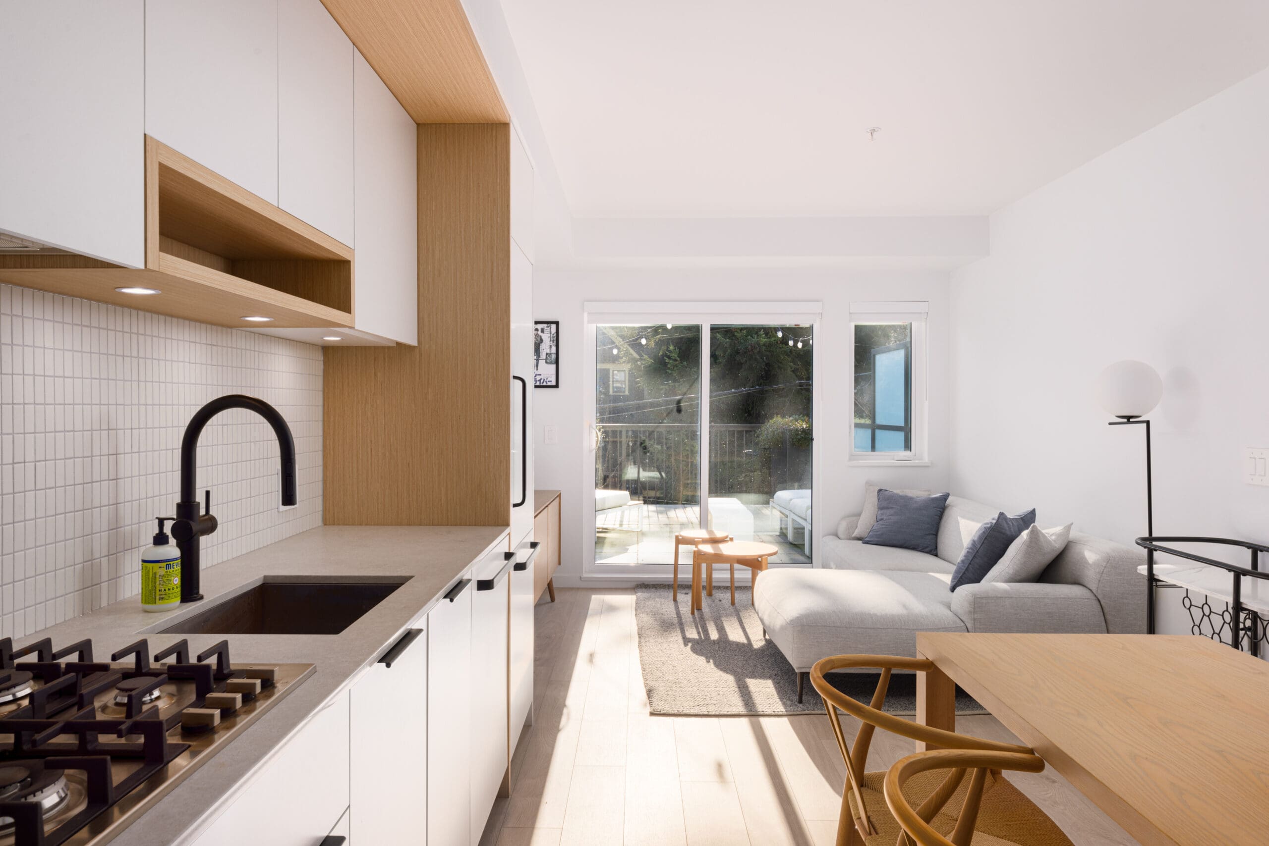 Modern kitchen and living area with white cabinets, a black faucet, gas stove, light wood accents, a gray sofa, wooden chairs, and large windows opening to a patio.