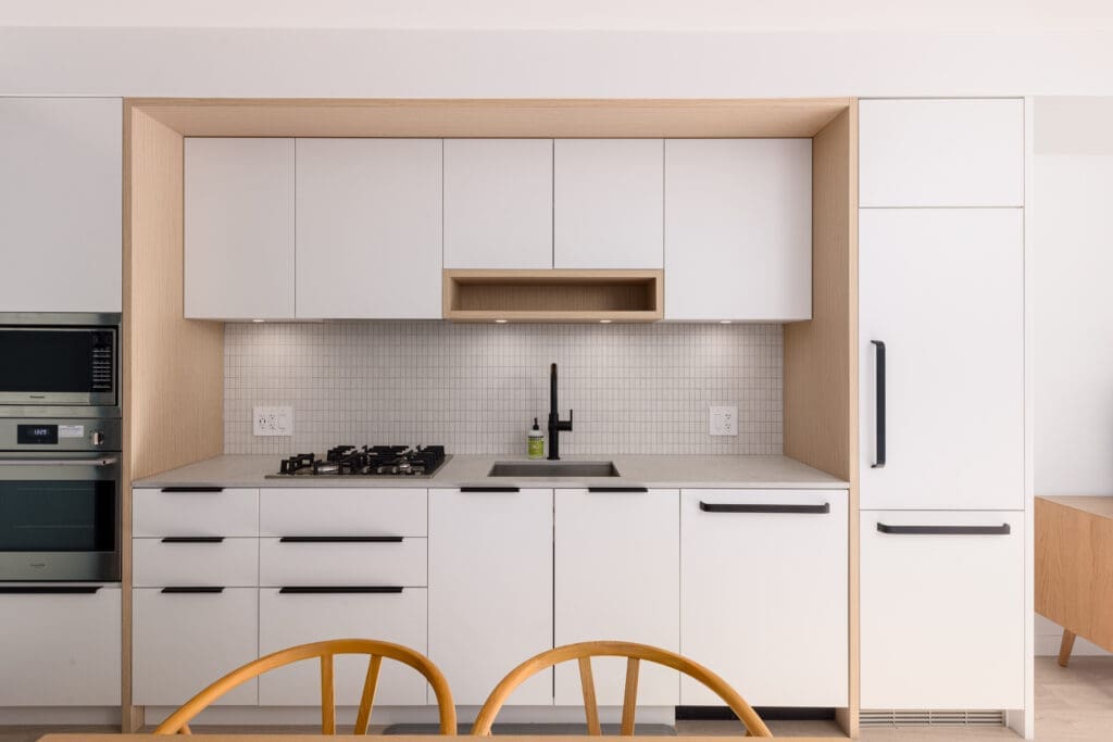Modern kitchen with white cabinets, black handles, a gas stove, sink, fridge, and oven. Two wooden chairs in the foreground.